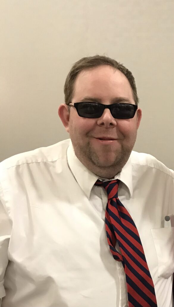 Wearing sunglasses, a smiling Kevin, host of the Troublemaker Experience, wears a white collared shirt with a diagonal striped tie.